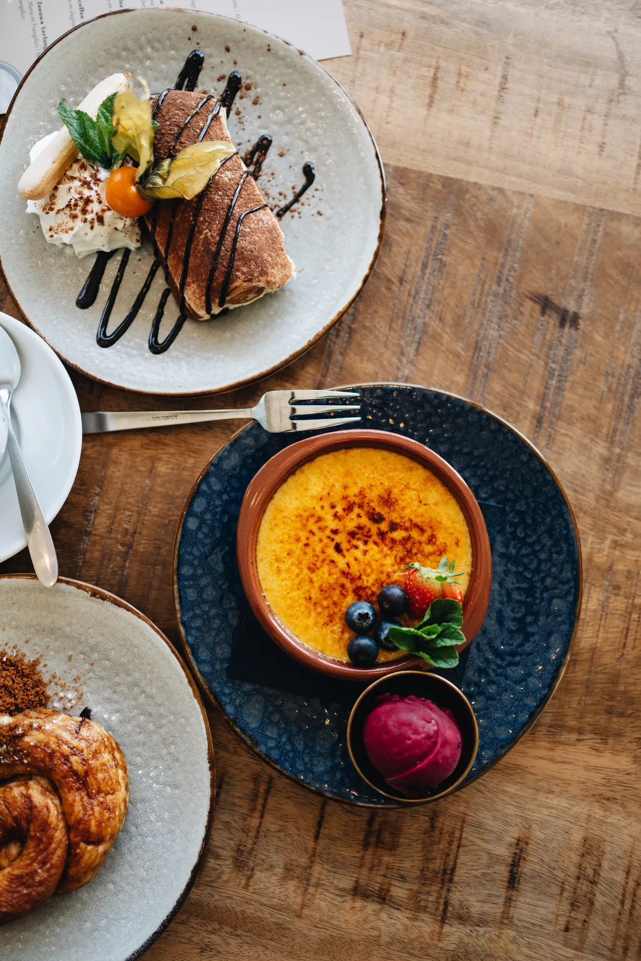 Close-up van verschillende nagerechten op een houten tafel, waaronder een schaaltje crème brûlée met bosbessen en frambozenijs, en een bord met tiramisu en chocoladesaus.